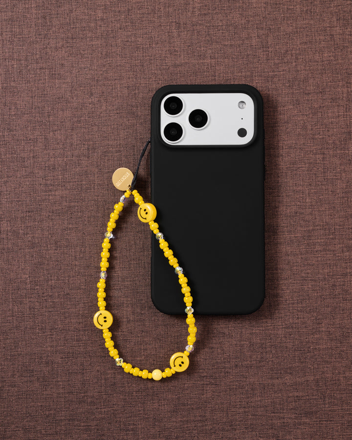 Yellow smiley bead phone strap looped onto a black phone case, its glossy beads and smiley faces bright against dark textured fabric.