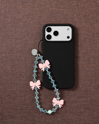 Beaded pink-bow strap looped onto a black phone case, pink bows and faceted ice-blue beads with a mint accent on brown fabric