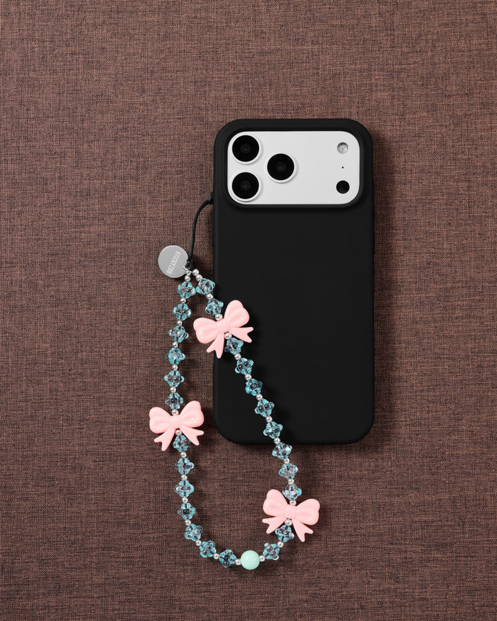Beaded pink-bow strap looped onto a black phone case, pink bows and faceted ice-blue beads with a mint accent on brown fabric