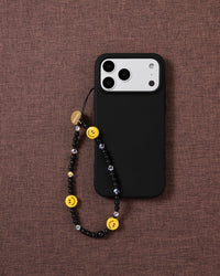 Black smiley beaded strap looped onto a black phone case on textured fabric, yellow smiley beads and faceted clear accents hanging in view.