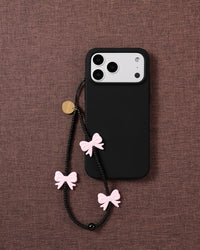 Midnight Bow strap on a black phone case over mauve fabric, pink bows and matte black beads hanging from the cord loop.