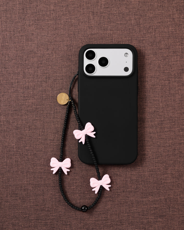 Midnight Bow strap on a black phone case over mauve fabric, pink bows and matte black beads hanging from the cord loop.