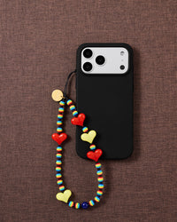 STICKYZOO Rainbow Heart strap looped onto a black phone case, red and yellow hearts and rainbow beads bright against brown fabric.