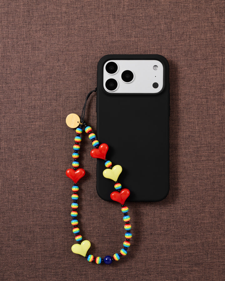 STICKYZOO Rainbow Heart strap looped onto a black phone case, red and yellow hearts and rainbow beads bright against brown fabric.