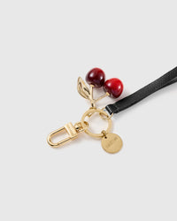 Macro close-up of the glossy red cherry charm on a gold-tone stem and leaf with gold clasp, ring and engraved STICKYZOO disc.