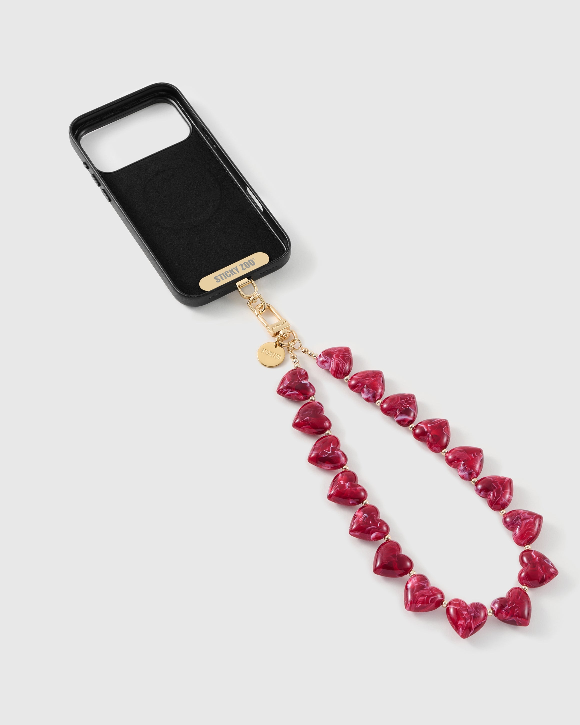Inside of a black phone case showing the MagSafe ring and gold STICKYZOO card connector, with ruby-red marbled heart beads on a gold hook.