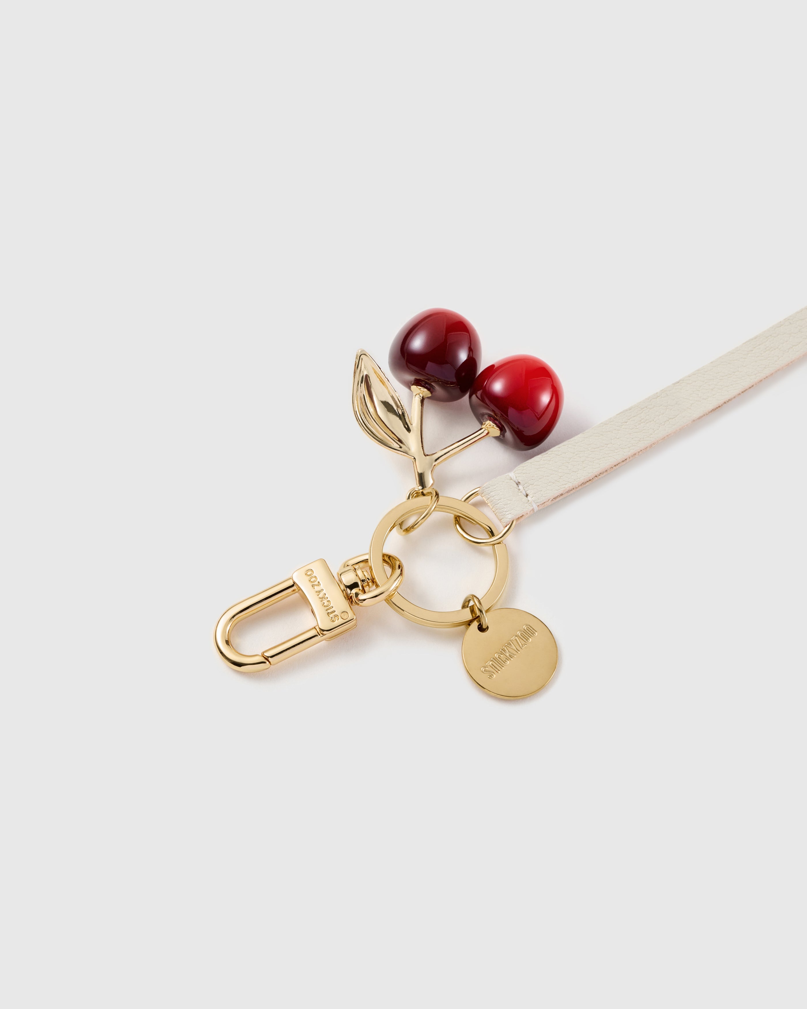 Macro of the STICKYZOO cherry charm: two glossy red cherries with a polished gold-tone leaf, above a gold ring and an engraved STICKYZOO disc.