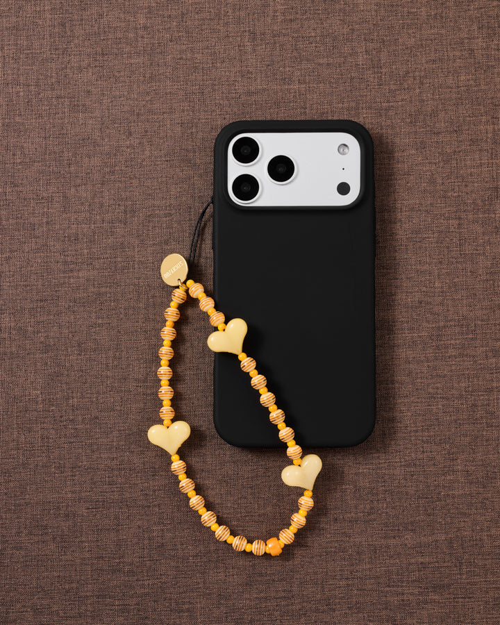 Apricot Heart beaded phone strap attached to a black phone case, showing three glossy yellow hearts and striped beads.