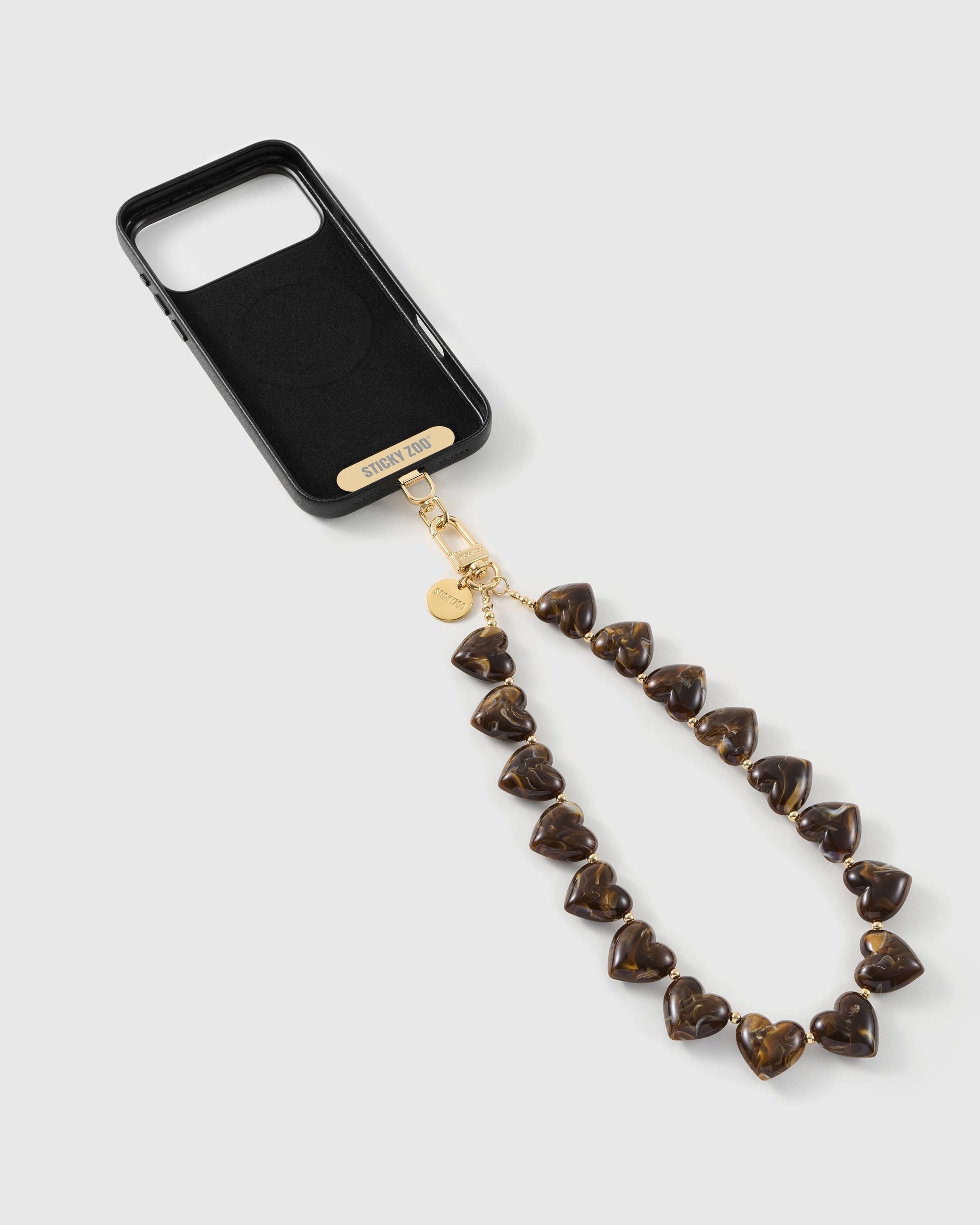 Amber-and-cocoa marbled heart beads draped from a black phone, with the gold-tone STICKYZOO strap card and engraved disc.