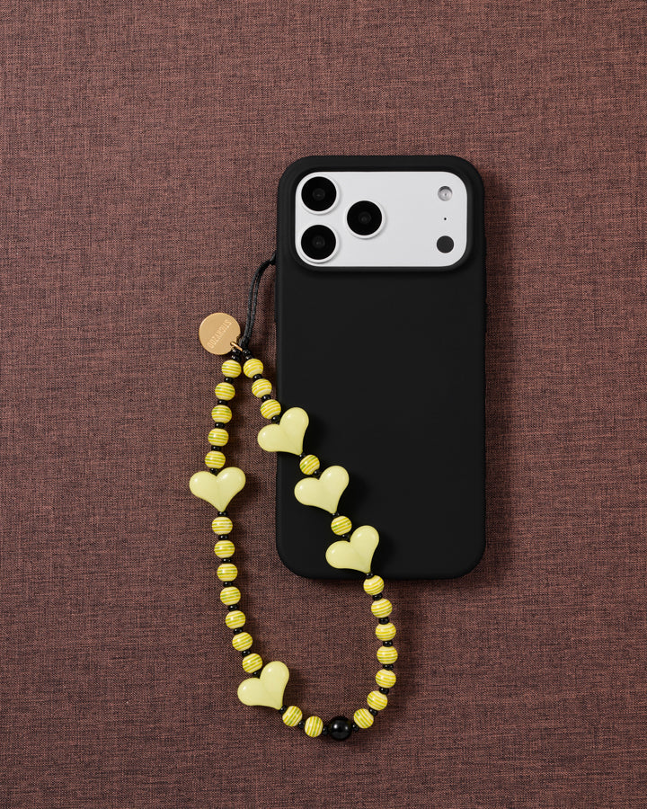 Lemon Heart beaded strap looped onto a black phone case on mauve fabric, pale yellow hearts and striped beads hanging free