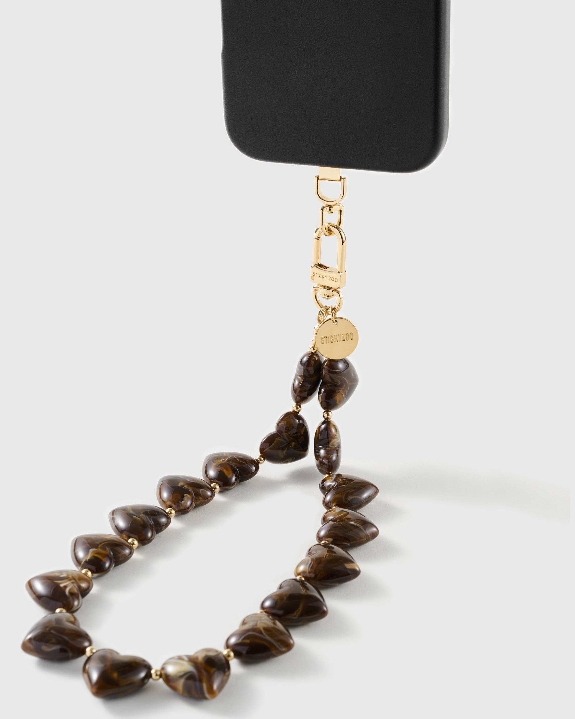 Tortoiseshell Heart strap on a black phone, showing the gold-tone STICKYZOO connector, engraved disc and glossy amber marbled heart beads.