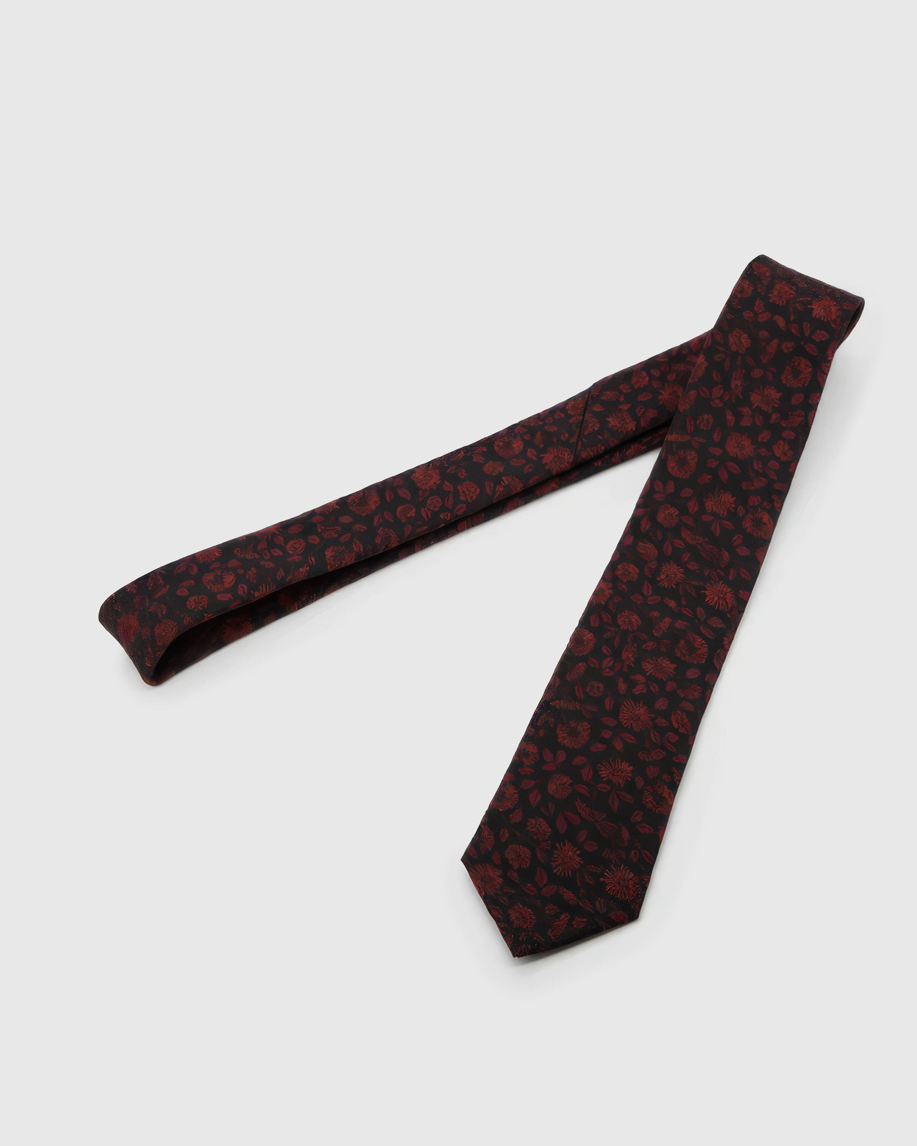 Noir Crimson Bloom Tie laid full length on white, showing a dark ground layered with crimson botanical blooms and shadowed leaves from blade to tail.