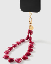 Black phone case corner held by the gold STICKYZOO clasp and engraved disc, above a strand of glossy ruby-red marbled heart beads.