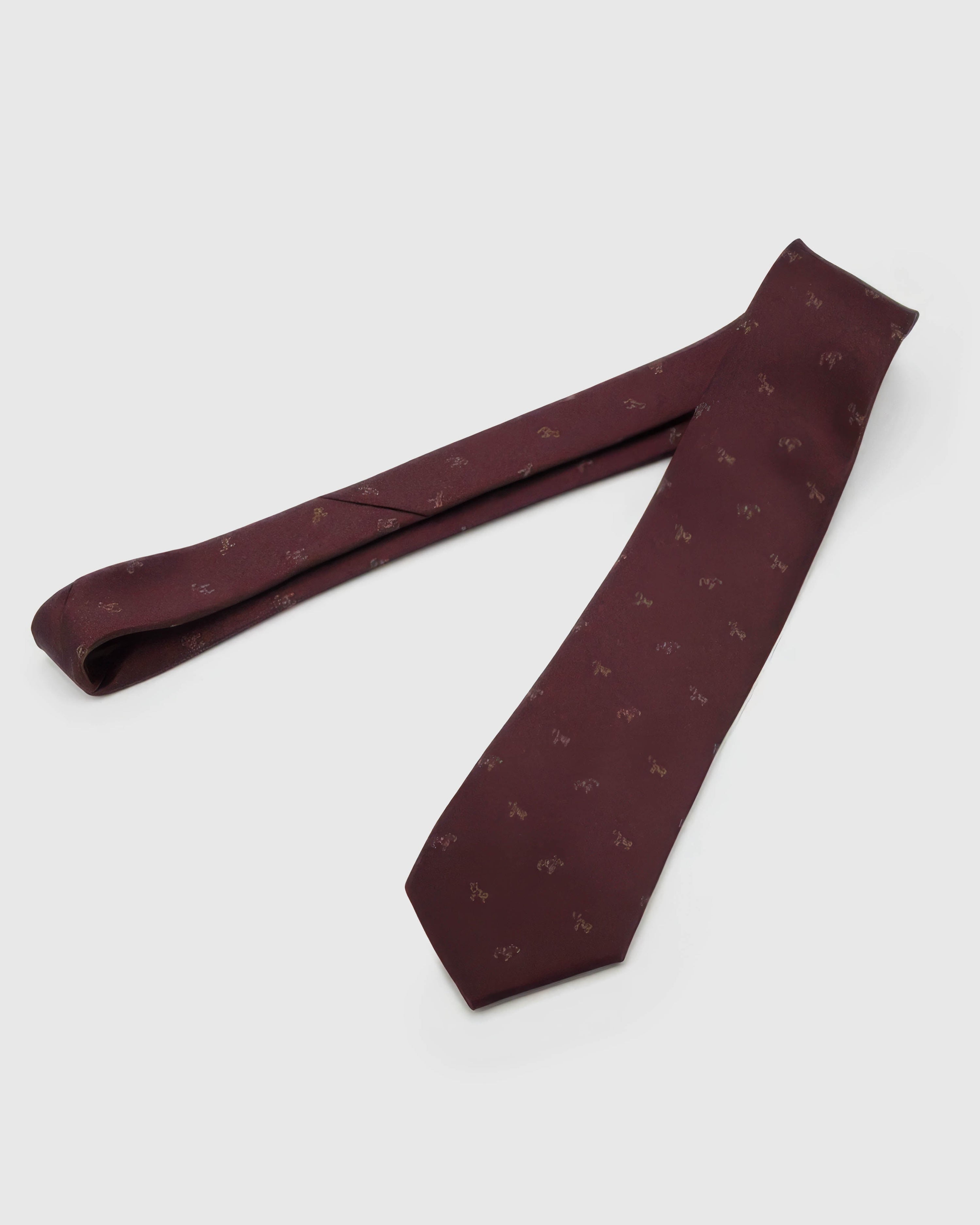 Burgundy Glyph Tie laid full length on white, showing a burgundy ground scattered with small muted-gold calligraphic glyphs from blade to tail.
