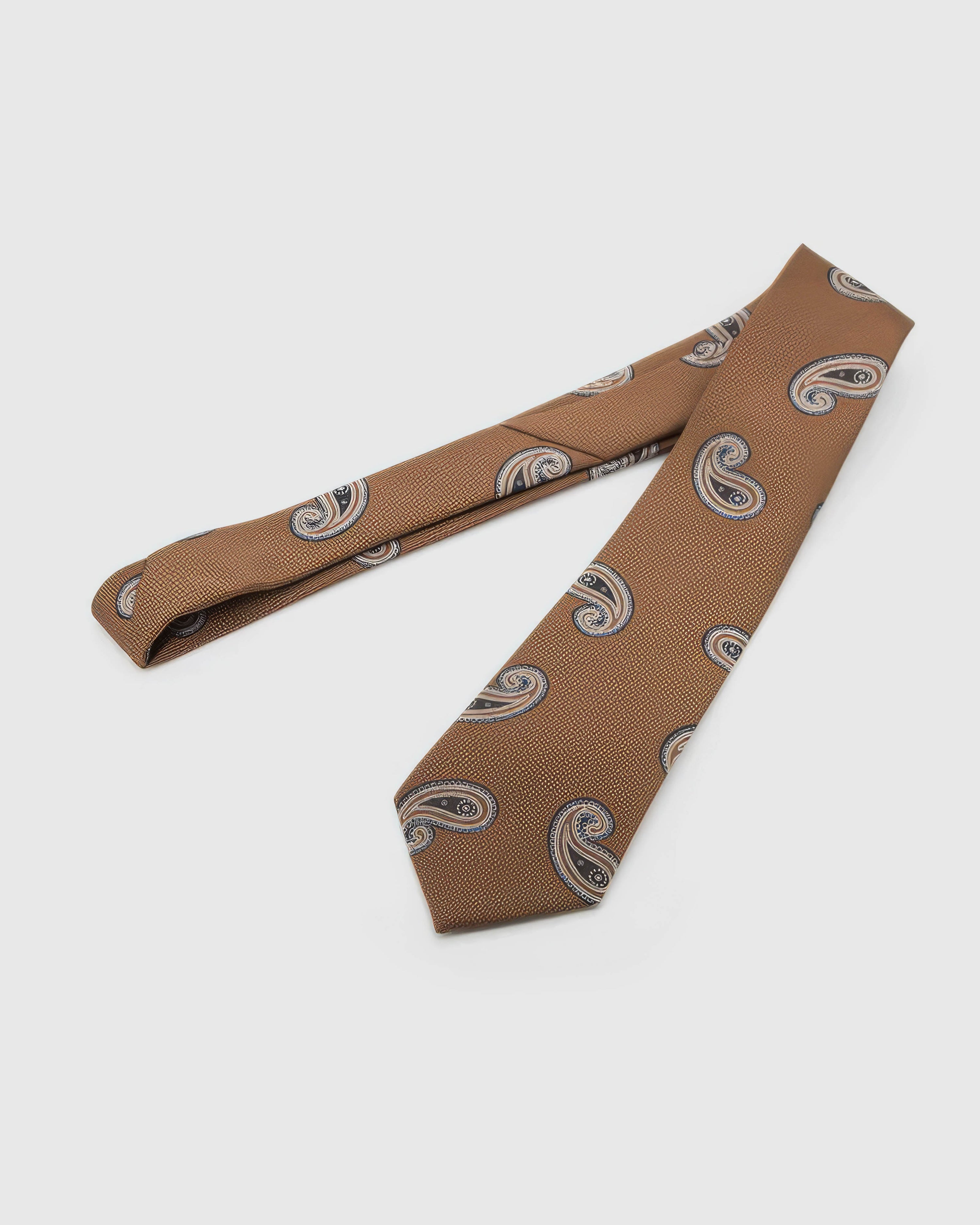 Cognac Paisley Tie laid full length on white, showing a cognac-toned ground with sweeping black, cream, and brown paisley forms from blade to tail.