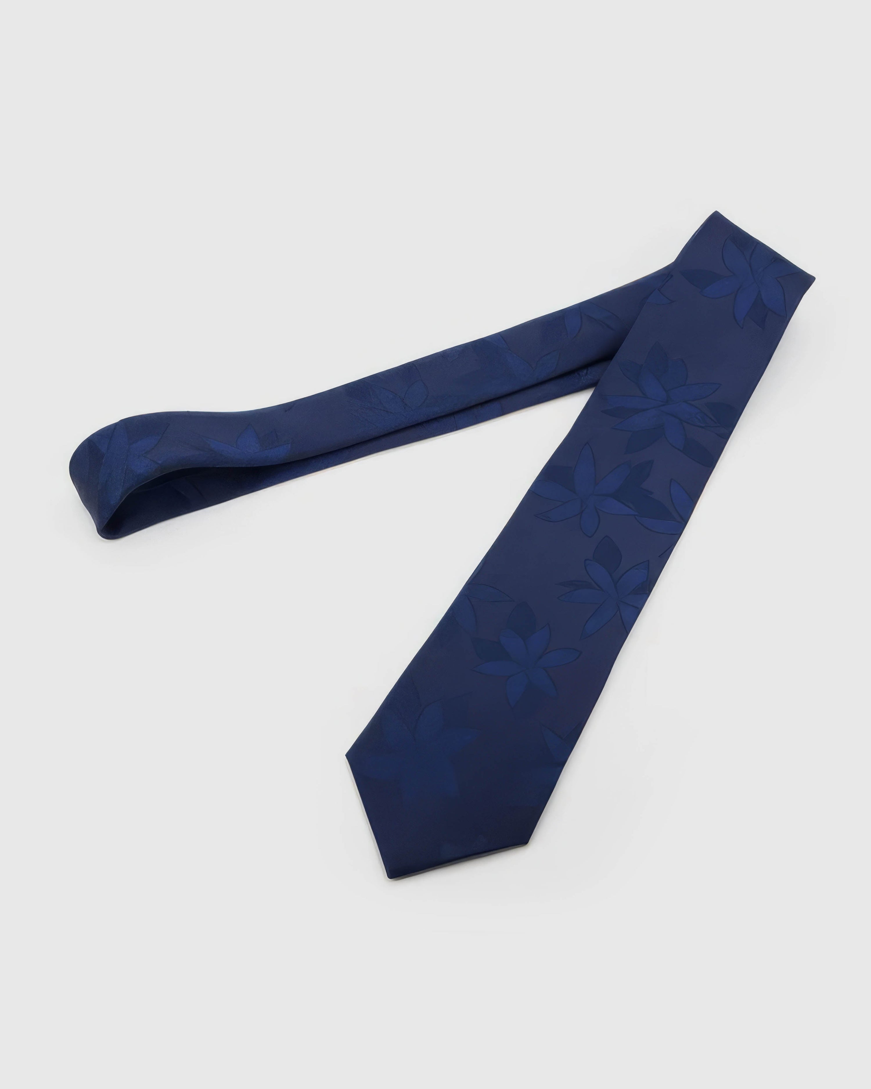 Sapphire Tonal Bloom Tie laid full length on white, showing a sapphire field with oversized tonal flower silhouettes and soft botanical forms from blade to tail.