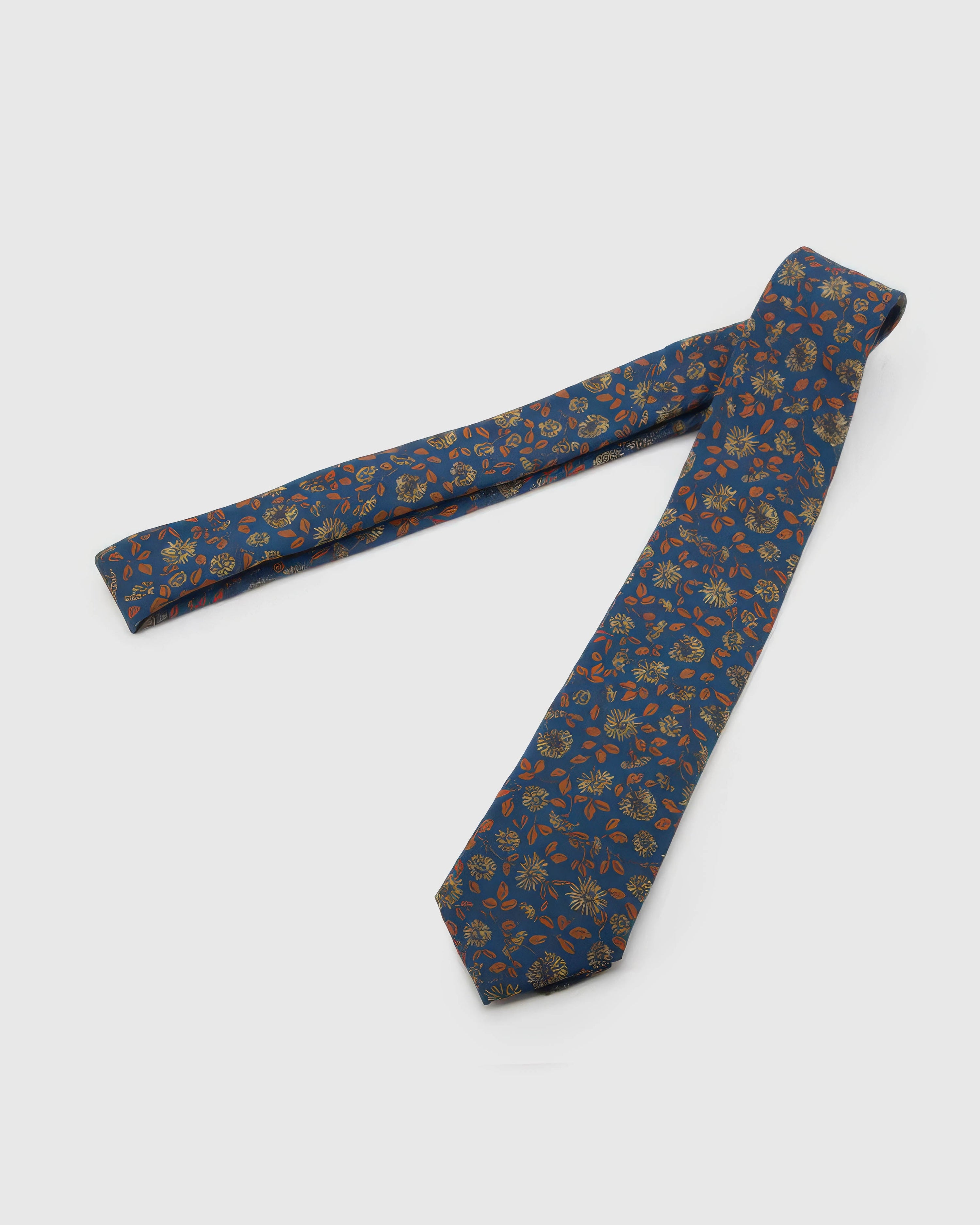 Cobalt Copper Bloom Tie laid full length on white, showing a cobalt ground with copper-toned leaves and pale-gold botanical blooms from blade to tail.