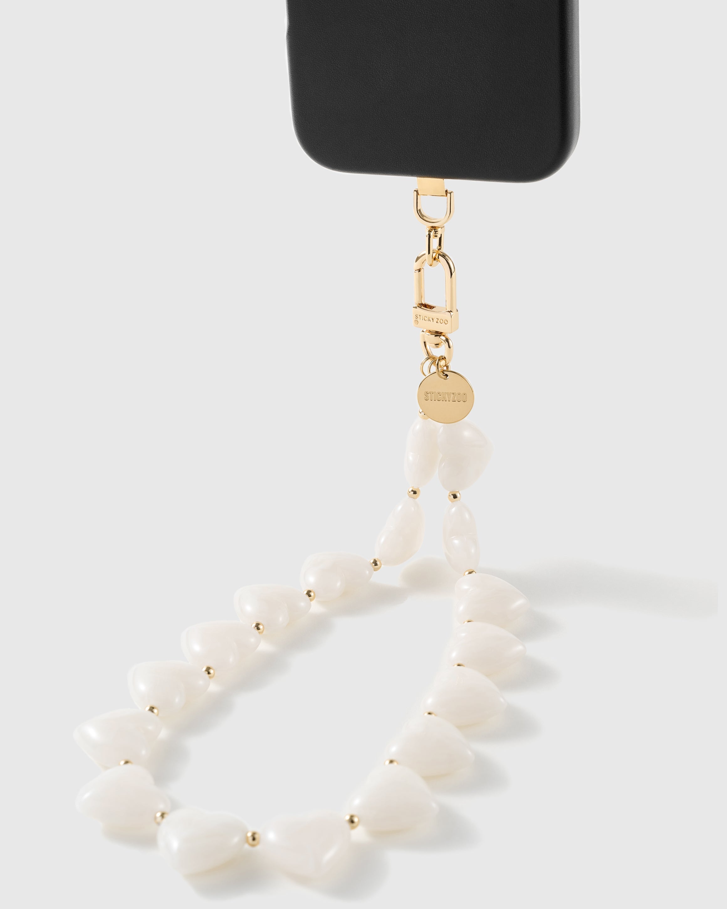 Milky white heart bead strap clipped to a black phone, showing the 18K gold-plated hook and STICKYZOO-engraved disc up close.
