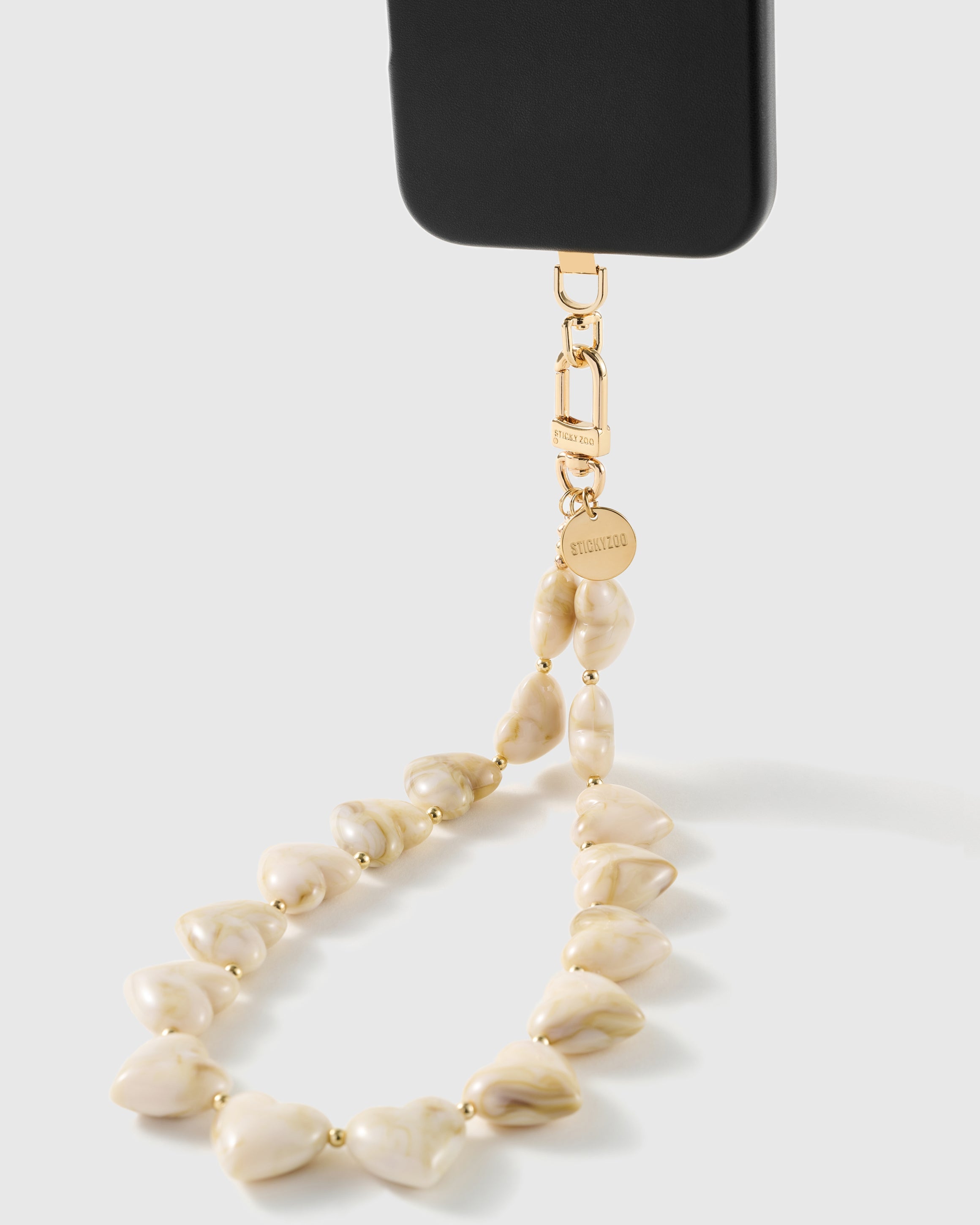 Marbled ivory heart bead strap clipped to a black phone case by a gold hook, connectors engraved STICKYZOO above a round gold disc.