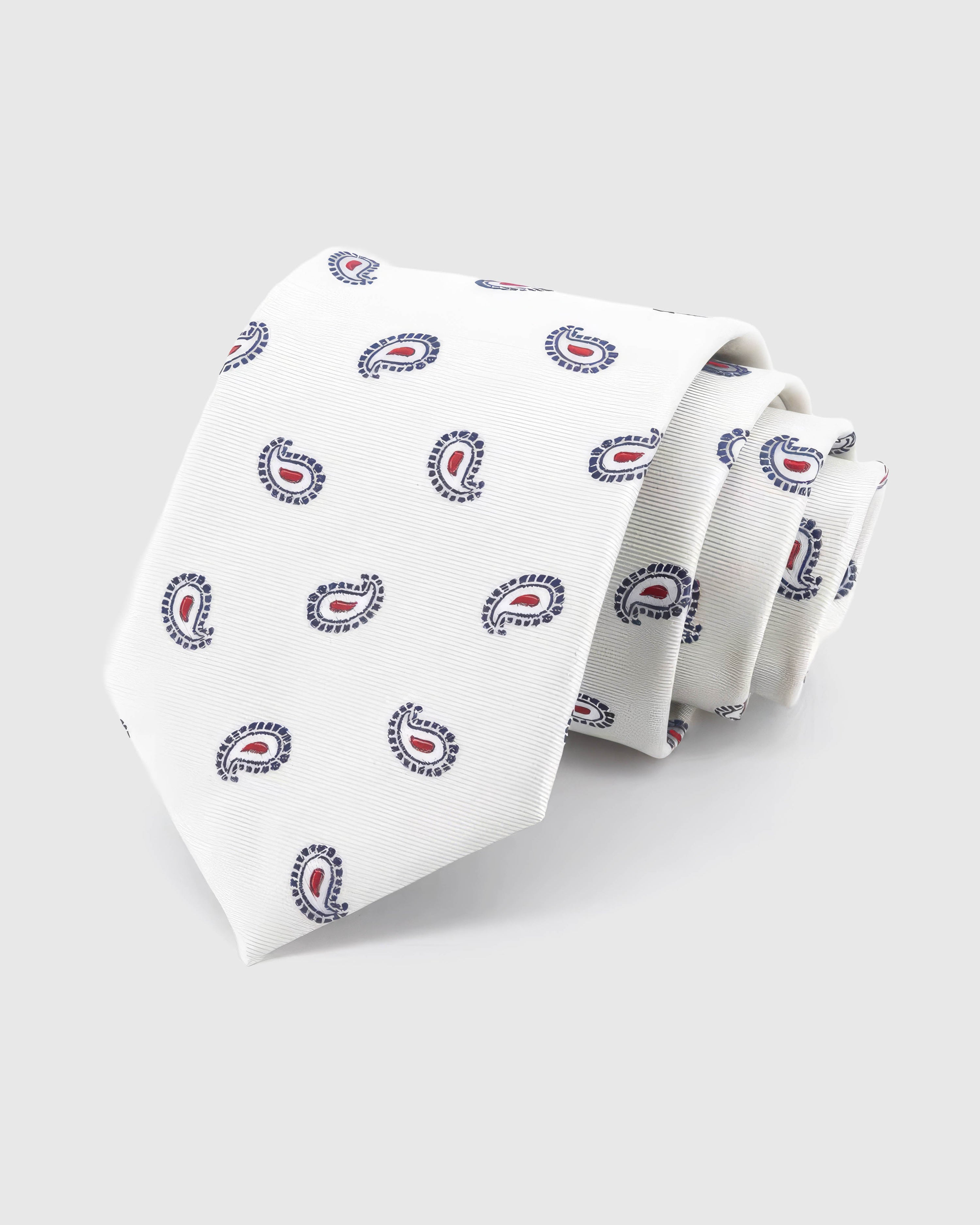 STICKYZOO Ivory Tricolor Paisley Tie rolled on white, showing an ivory ground with small navy-outline paisleys and crisp crimson centres across the front blade.