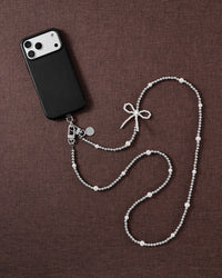 Silver-tone and faux-pearl bow cross-body phone strap styled beside a black phone on brown linen.