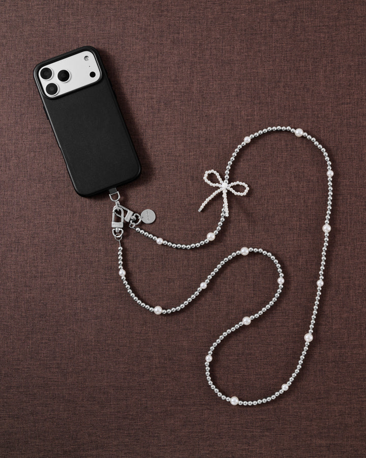 Silver-tone and faux-pearl bow cross-body phone strap styled beside a black phone on brown linen.
