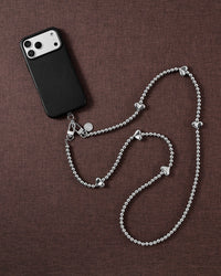 Silver-tone puffy-heart cross-body phone strap arranged beside a black phone case on brown fabric.
