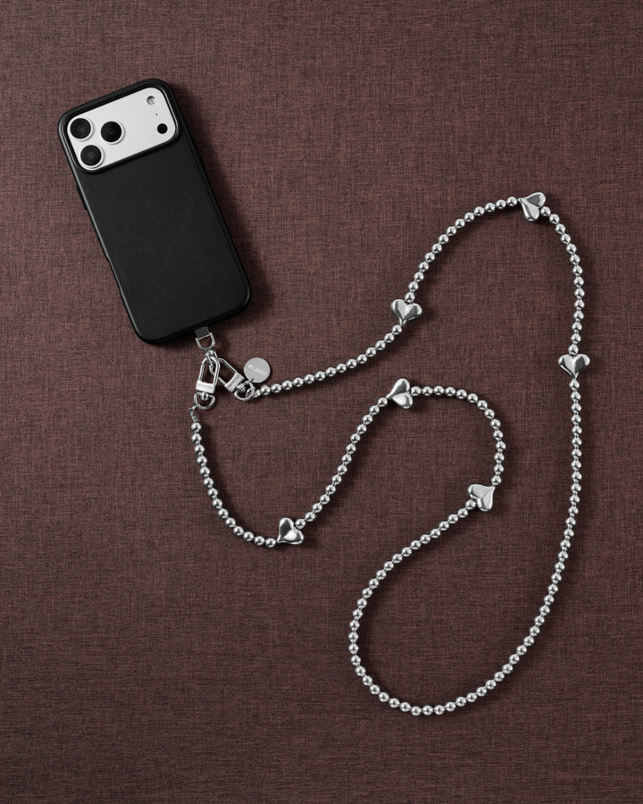 Silver-tone puffy-heart cross-body phone strap arranged beside a black phone case on brown fabric.