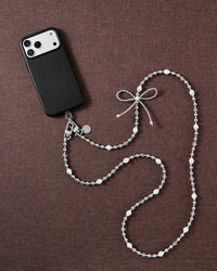 Faux-pearl and silver-tone bow cross-body phone strap styled beside a black phone on brown fabric.
