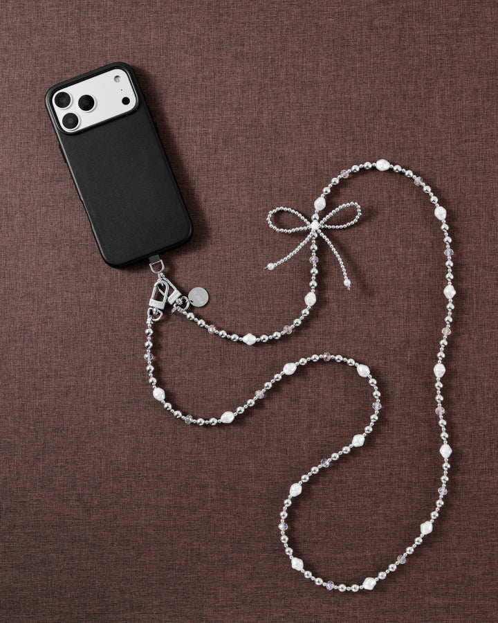Faux-pearl and silver-tone bow cross-body phone strap styled beside a black phone on brown fabric.