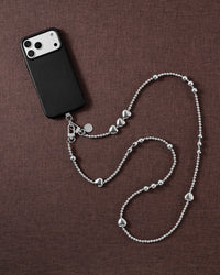 Silver-tone heart and bead cross-body phone strap styled beside a black phone case on brown linen.