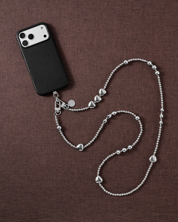 Silver-tone heart and bead cross-body phone strap styled beside a black phone case on brown linen.
