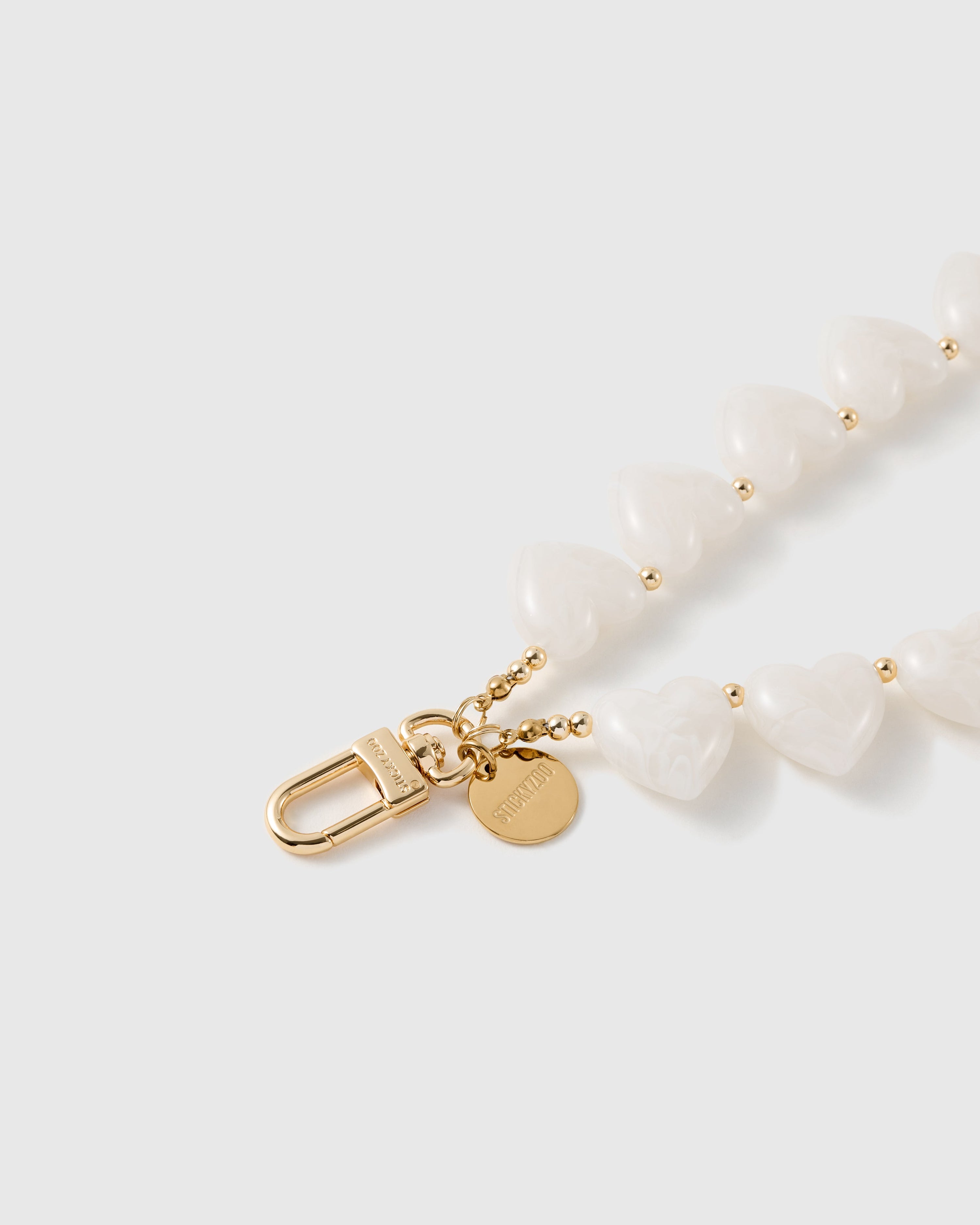 Close-up of the gold oval clasp and round STICKYZOO disc beside glossy milky white heart beads and small gold ball spacers.
