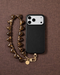Flat-lay of the Tortoiseshell Heart phone strap on brown linen: marbled amber heart beads, gold-tone clasp and engraved disc beside a black case.