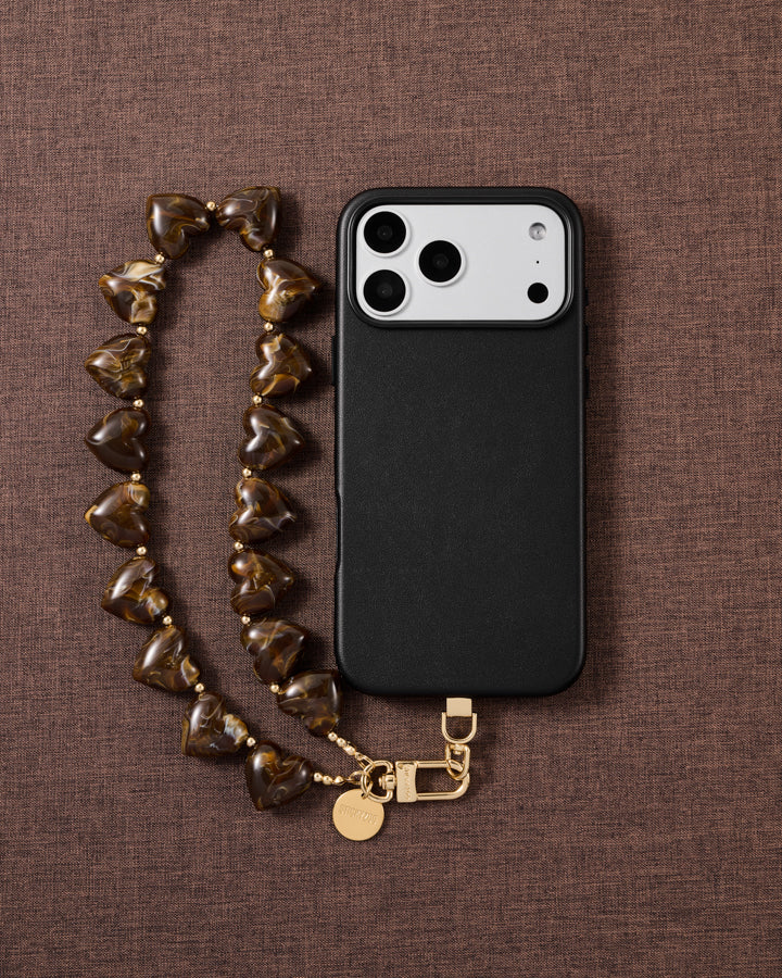 Flat-lay of the Tortoiseshell Heart phone strap on brown linen: marbled amber heart beads, gold-tone clasp and engraved disc beside a black case.