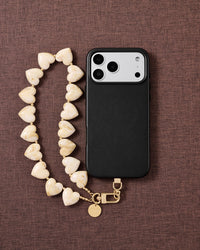 Ivory marbled heart bead strap looped beside a black phone case on brown fabric, gold hook and engraved disc resting at the base.