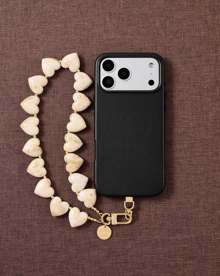 Ivory marbled heart bead strap looped beside a black phone case on brown fabric, gold hook and engraved disc resting at the base.