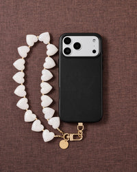 Flat-lay on mauve fabric: a black phone beside the milky white heart bead strand with gold clasp and engraved disc.