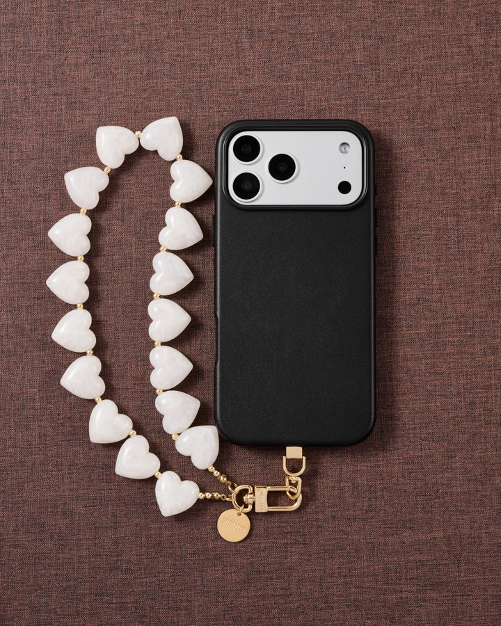 Flat-lay on mauve fabric: a black phone beside the milky white heart bead strand with gold clasp and engraved disc.