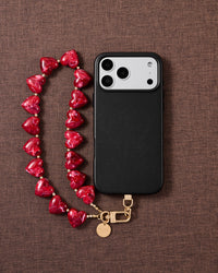 Ruby-red marbled heart bead strap laid on brown textured fabric beside a black phone case, with gold-plated hook and engraved disc.