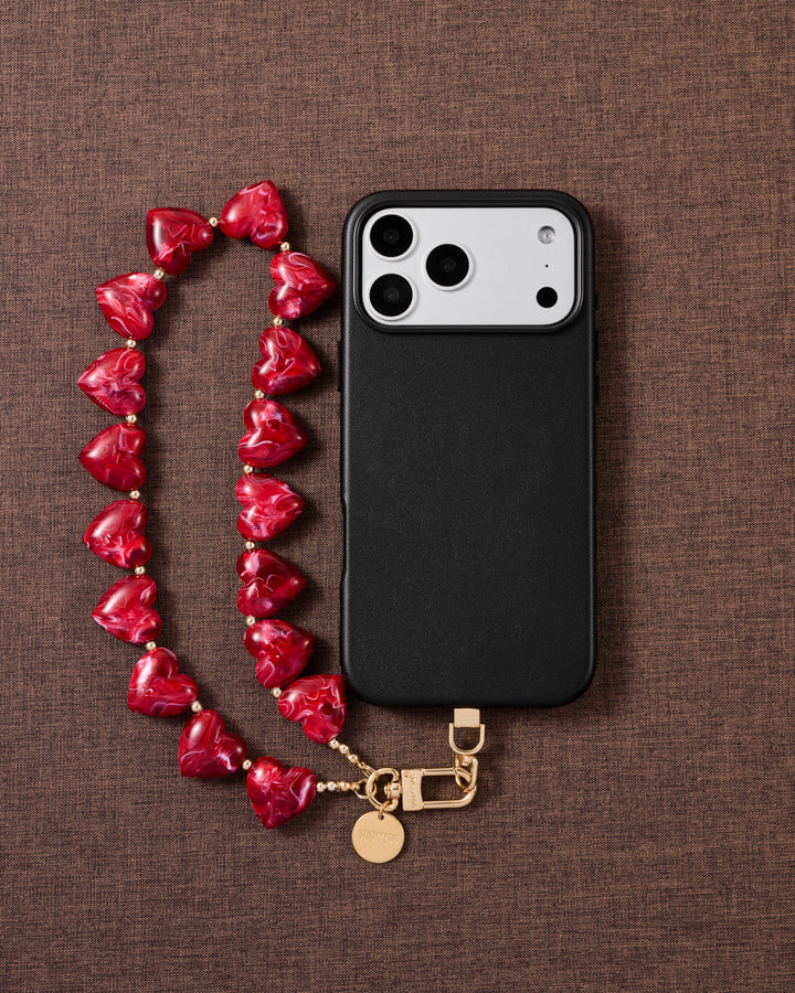 Ruby-red marbled heart bead strap laid on brown textured fabric beside a black phone case, with gold-plated hook and engraved disc.